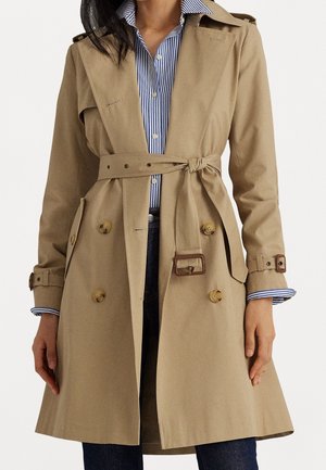 Femme portant un trench-coat beige à double boutonnage avec ceinture nouée à la taille, sur une chemise à rayures bleues et blanches à boutons, et un pantalon foncé.