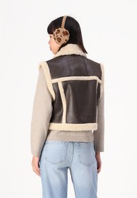 Brun faux læder vest med beige shearling kant, der har en struktureret design og detaljer på rygpanelet. Letvægts striktrøje og blå jeans.