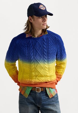 Knit sweater featuring a gradient from blue to yellow, with cable patterns. Worn over a multicolored plaid shirt and denim jeans.