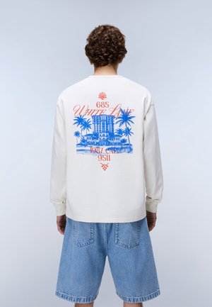 Muž s kudrnatými vlasy nosící bílou košili s dlouhým rukávem s modrým a oranžovým tropickým motivem budovy a palmy, doplněnou volnými džínovými šortkami.