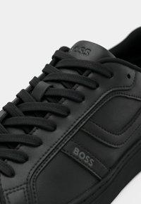 Svarte skinn-sneakers med teksturert overflate, flate svarte lisser og subtil logo på siden. Rund tå og polstret krage.