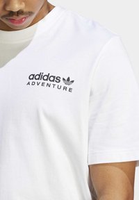 Camiseta de algodón blanca con cuello redondo y mangas cortas; texto negro serigrafiado "adidas ADVENTURE" junto a un logotipo.