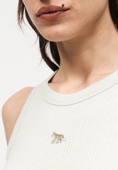 Κοντινό πλάνο στο κάτω μέρος του προσώπου και τον λαιμό ενός ατόμου που φοράει λευκό αμάνικο ραβδωτό τοπ με μια μικρή κεντημένη σκύλο στο στήθος.