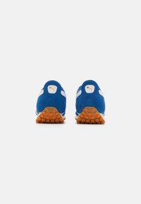 Zapatillas deportivas de ante azul con acentos de tela blanca y suela de goma naranja con clavos circulares para tracción. Detalle del logo dorado en el talón.