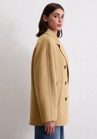 Double-breasted coat in light beige wool, featuring wide lapels, two front pockets, and brown buttons, styled with relaxed shoulders.