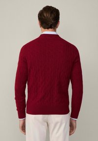 Roter Kabelstrickpullover mit strukturiertem Muster, gerippten Bündchen und Saum sowie Rundhalsausschnitt. Über einem hellblauen Hemd mit Kragen getragen.