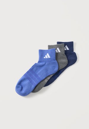 Trois paires de chaussettes basses en bleu, gris et marine, chacune avec un logo blanc sur le bord côtelé, disposées en chevauchement sur un fond clair.