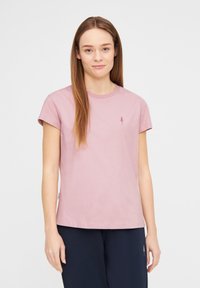 Rosa Baumwoll-T-Shirt mit kurzen Ärmeln, rundem Ausschnitt und kleiner Baumstickerei auf der Brust. Getragen mit dunklen Hosen.