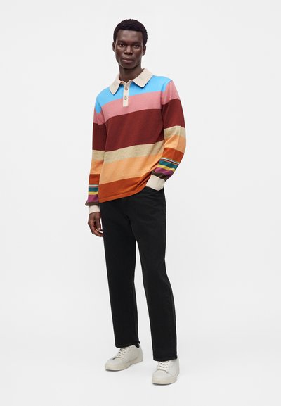 Homme debout portant un polo à rayures multicolores à manches longues, un pantalon noir et des baskets blanches sur un fond blanc uni.