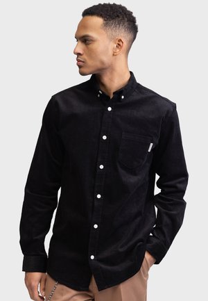 Black corduroy button-up shirt with a pocket, white buttons, and a collared neckline, featuring a soft texture and tailored fit.