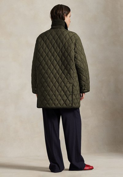 Inimene seisab selg nähtaval, seljas oliivrohelise tikandiga jakk, lahedad tumedad püksid ja punased kingad neutraalse taustaga.