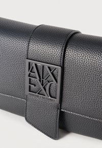 Μαύρο πορτοφόλι από δέρμα με υφή και μεταλλικό κούμπωμα με το λογότυπο Armani Exchange σε ανοιχτό φόντο.