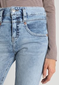 Světlé modré džíny z denimu s vysokým pasem, designem na pět kapes a měděnými akcenty. Mají mírné vyblednutí a přiléhavý siluetu.