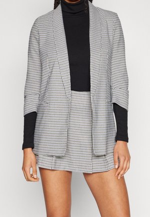 Vrouw draagt een zwarte coltrui met een zwart-witte pied-de-poule blazer en bijpassende rok, staand tegen een lichte achtergrond.