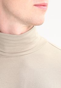Beige Rollkragenoberteil aus geripptem Stoff. Verfügt über einen hohen Kragen und eine subtile Textur, aus der Perspektive des Halsausschnitts nach oben gezeigt.