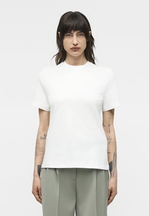 Mujer con cabello oscuro y tatuajes que lleva una camiseta blanca de manga corta lisa y pantalones gris verdosos claros, de pie contra un fondo gris.