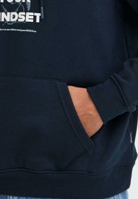 Temno modra majica s kapuco z mehkim teksturo, ki ima sprednji žep in bel grafični napis. Robovi na manšetah in spodnjem delu zagotavljajo prileganje.
