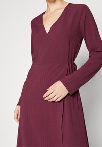 Kvinna iklädd en långärmad, djup V-ringad, ribbad plommonfärgad omslagsklänning knuten i midjan, med en hand vilande på kjolen.