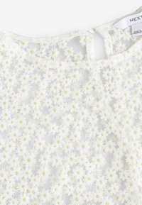 Top blanc avec un design brodé de fleurs comprenant de petites marguerites et des accents dorés. Matière en maille texturée et fermeture à bouton à l'arrière.