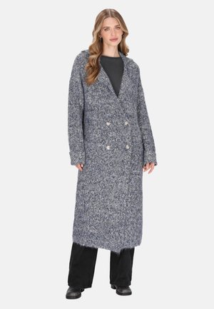 Frau mit langen, welligen Haaren, die einen langen, grauen, doppelt geknöpften Strickcardigan über schwarzen Hosen und schwarzen Stiefeln trägt und vor einem weißen Hintergrund steht.