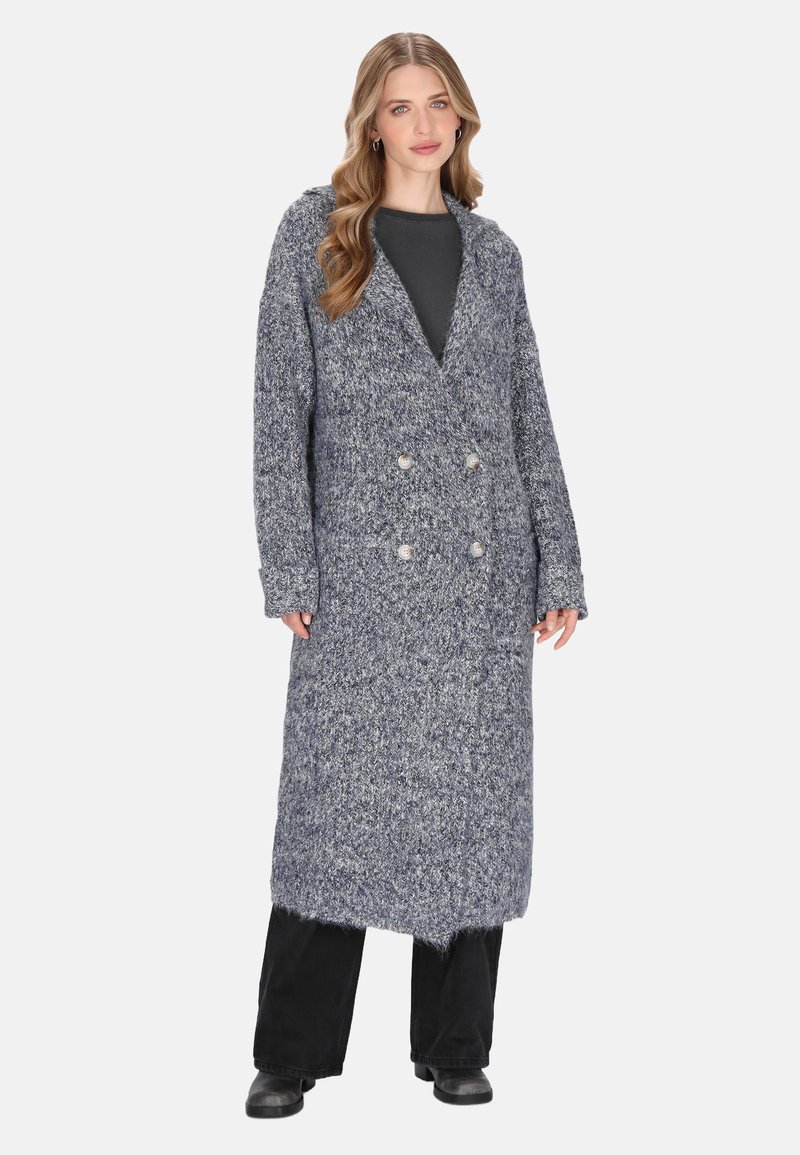 Donna con lunghi capelli mossi che indossa un lungo cardigan grigio a doppio petto sopra pantaloni neri e stivali neri, in piedi contro uno sfondo bianco.