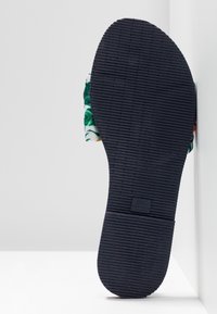 Sandalens sula har texturerad svart gummi och en slät, rundad form. Synliga bladmönster från det övre materialet vid kanterna.