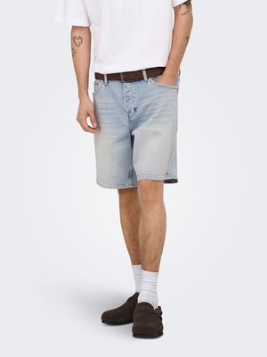 Personne portant un short en jean bleu clair, un t-shirt blanc, des chaussettes blanches, des chaussures marron sans lacets et une ceinture marron, avec des tatouages visibles sur les bras.