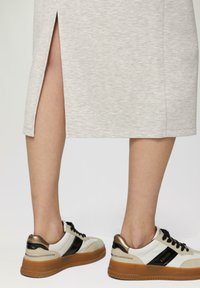Grauer, knielanger Rock mit seitlichem Schlitz, kombiniert mit weißen und schwarzen Sneakers mit goldenen Akzenten und strukturierten Sohlen.