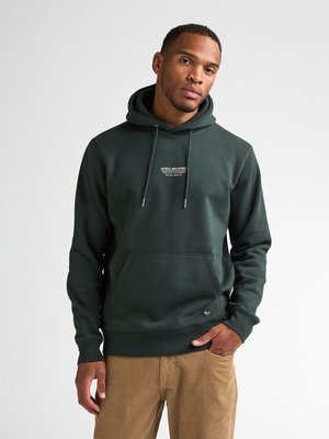 Man wearing a dark green hoodie with front pocket and beige corduroy pants, standing against a plain light background.