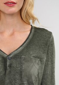 Olivenfarbener Langarmtop mit V-Ausschnitt, ausgestattet mit einer Brusttasche und einem weichen, leicht strukturierten Stoff in entspannter Passform.