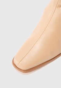 Beige bőr bokacsizma hegyes orral, látható varrással és lapos barna talppal. Simább textúra, minimalista design.