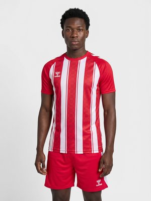 Ung mand står iført en rød- og hvidstribet sportstrøje og røde shorts med brandlogo på brystet og shorts.