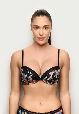 Frau mit dunklen Haaren, im Pferdeschwanz, trägt einen schwarzen BH mit Blumenmuster und Spitzenbesatz, blickt nach vorne vor einem schlichten hellen Hintergrund.