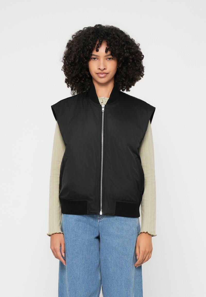 Casaco bomber preto sem mangas com fecho éclair, bainha canelada e textura suave, usado sobre uma blusa de manga longa verde-clara e jeans azuis.