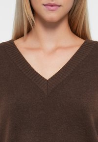 Brun, v-hals genser med strukturert strikk. Stoffet ser mykt ut, med ribbestrikket hals og en generelt avslappet passform.