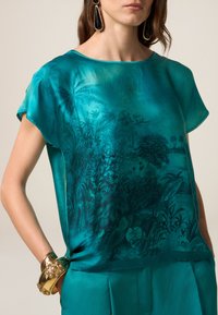 Blusa di seta turchese con stampa di piante tropicali, maniche corte, scollatura ampia e vestibilità rilassata. Abbinata a un bracciale in oro con texture.