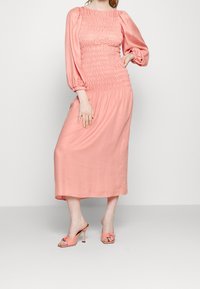 Vaaleanpunainen polvipituinen mekko, jossa on kerrattu yläosa ja puffihihat; satiinimainen pinta. Yhdistetty vaaleanpunaisiin avonaisiin sandaaleihin, joissa on nauhakiinnitys.