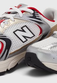 Baskets New Balance blanches, rouges et grises avec des panneaux en mesh, des lacets et le logo distinctif « N » sur le côté et la semelle.