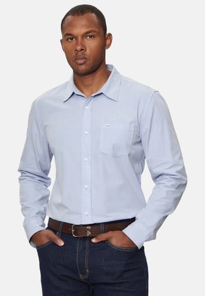 Camisa azul claro de manga larga y botones, con un bolsillo en el pecho, hecha de una tela suave; combinada con jeans azul oscuro y un cinturón marrón.