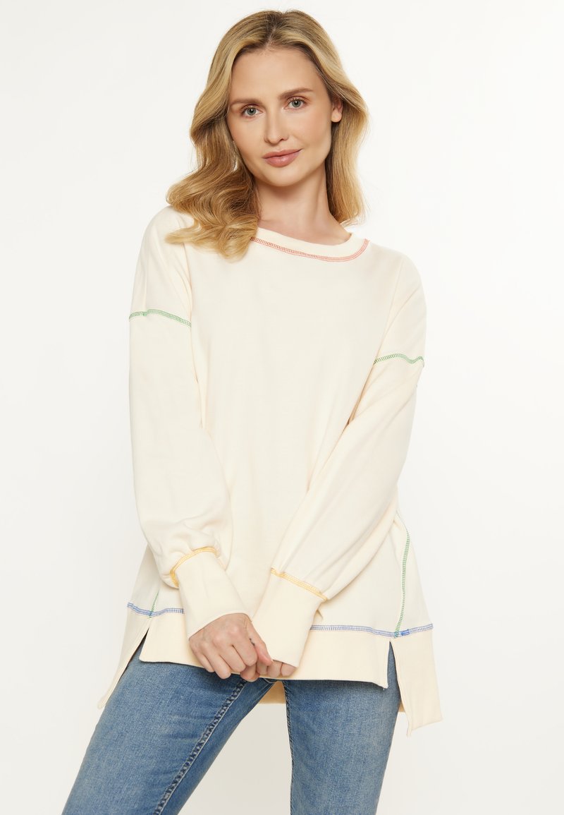 Maglione color crema chiaro con maniche lunghe, caratterizzato da cuciture multicolori in rosa, verde e blu. Le fessure laterali aumentano il comfort.