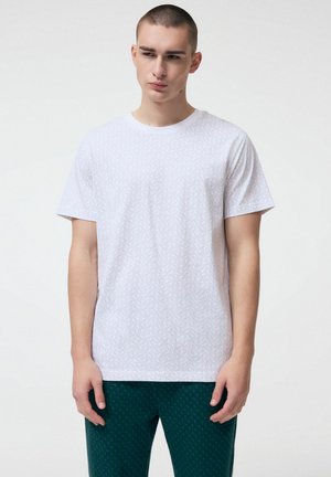 Młody mężczyzna z krótkimi włosami, ubrany w białą koszulkę w wzory i ciemnozielone spodnie, stoi na tle gładkiego, jasnego tła.