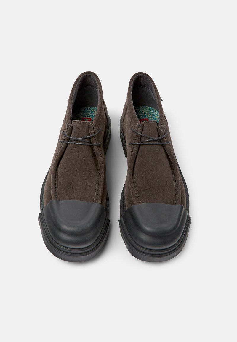 Braune Wildlederschuhe mit einer schwarzen Gummikappe, minimaler Schnürung und strukturierten Innensohlen. Das Design verfügt über eine abgerundete Form für zusätzlichen Komfort.