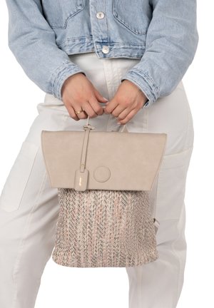 Personne en veste en jean et pantalon blanc tenant un sac à dos beige tissé avec rabat et poignée supérieure devant un fond blanc.