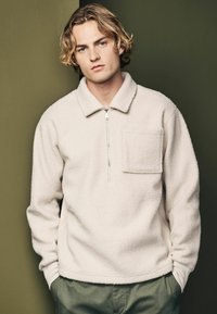Pulover din fleece de culoare albă, cu fermoar pe jumătate în față, guler și un buzunar pe piept. Textură moale cu manșete și tiv din tricot.