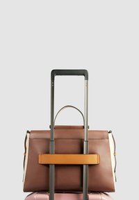 Braune Lederhandtasche mit hellbraunen Akzenten, oben auf einem rosa Koffer befestigt, mit einem strukturierten Design und zwei Griffen.