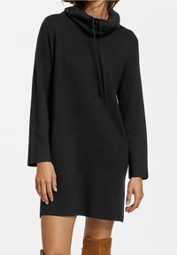 Robe noire à col bénitier avec des manches longues, en tissu doux, coupe décontractée et fentes sur les côtés ; associée à des bottes en daim marron hauteur genoux.