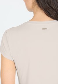 Helles beige Kurzarmhemd mit glatter Textur und einem kleinen metallischen Logo-Tag auf der Rückseite. Einfaches, sauberes Design mit minimalen Nähten.