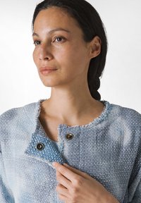 Giacca tessuta blu chiaro con bordi sfilacciati, bottoni metallici e superficie texturizzata. Il tessuto presenta un effetto sfumato e motivi sottili.