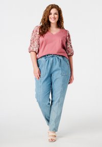Roze top met gedessineerde ballonmouwen; lichtblauwe denim broek met losse pasvorm, elastische tailleband en zijzakken. Open teen beige sandalen.