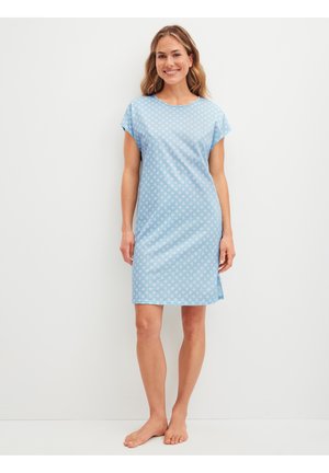 Vrouw staat op blote voeten, glimlachend, draagt een lichtblauwe jurk met korte mouwen tot op de knie met een subtiel stippenpatroon.